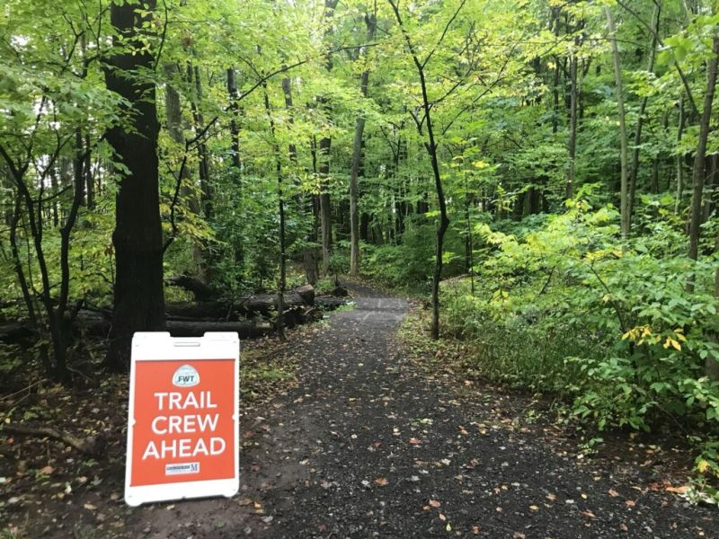 A footpath through a lush green forest with a large sign from Friends of Webster Trails reading "Trail Crew Ahead"