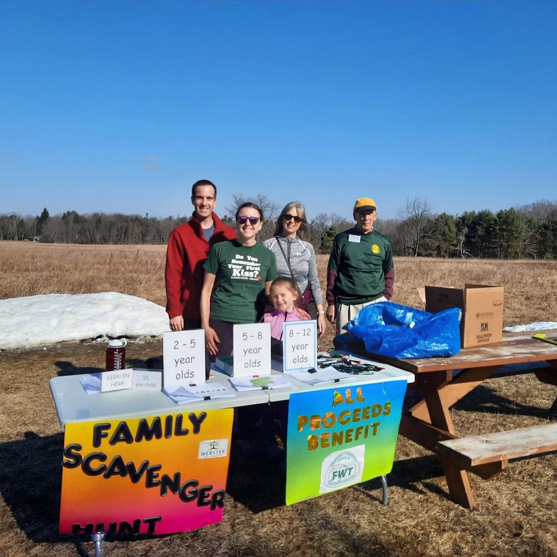 Four adults and one child behind a table with signs for a scavenger hunt. Blue skies and a brown field are in the background.