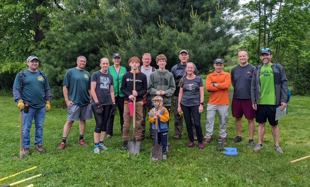A group of adults and one small child in front of evergreen trees. Many are holding shovels.